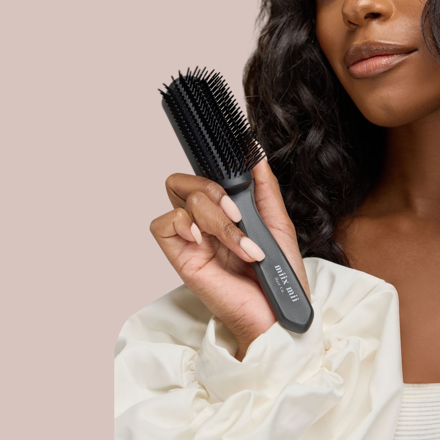 A confident woman with radiant brown skin and glossy, wavy hair holds a high-quality curl defining brush with firm yet flexible bristles. The sleek black handle, embossed with "Miix Mii Hair Co.," offers a comfortable grip, making it an essential tool for enhancing curl definition and detangling textured hair. Her poised expression and elegant cream-colored blouse add a touch of sophistication to the image.