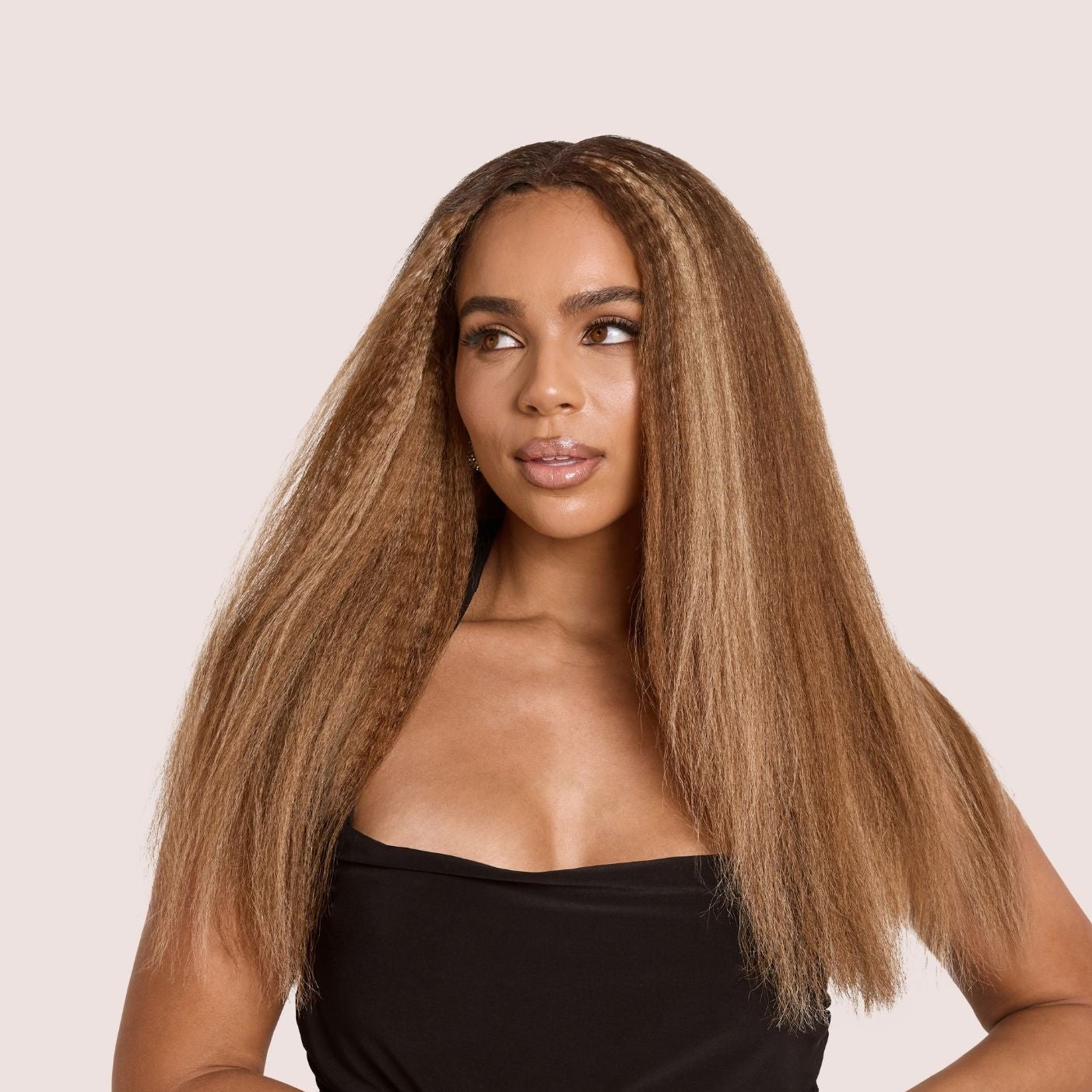 Confident woman with voluminous honey-blonde kinky straight hair extensions, styled with a luxurious texture that mimics natural blown-out hair. She poses in a sleek black dress, highlighting the fullness and movement of the premium luxury textured hair extensions, perfect for adding length and volume with a natural look.