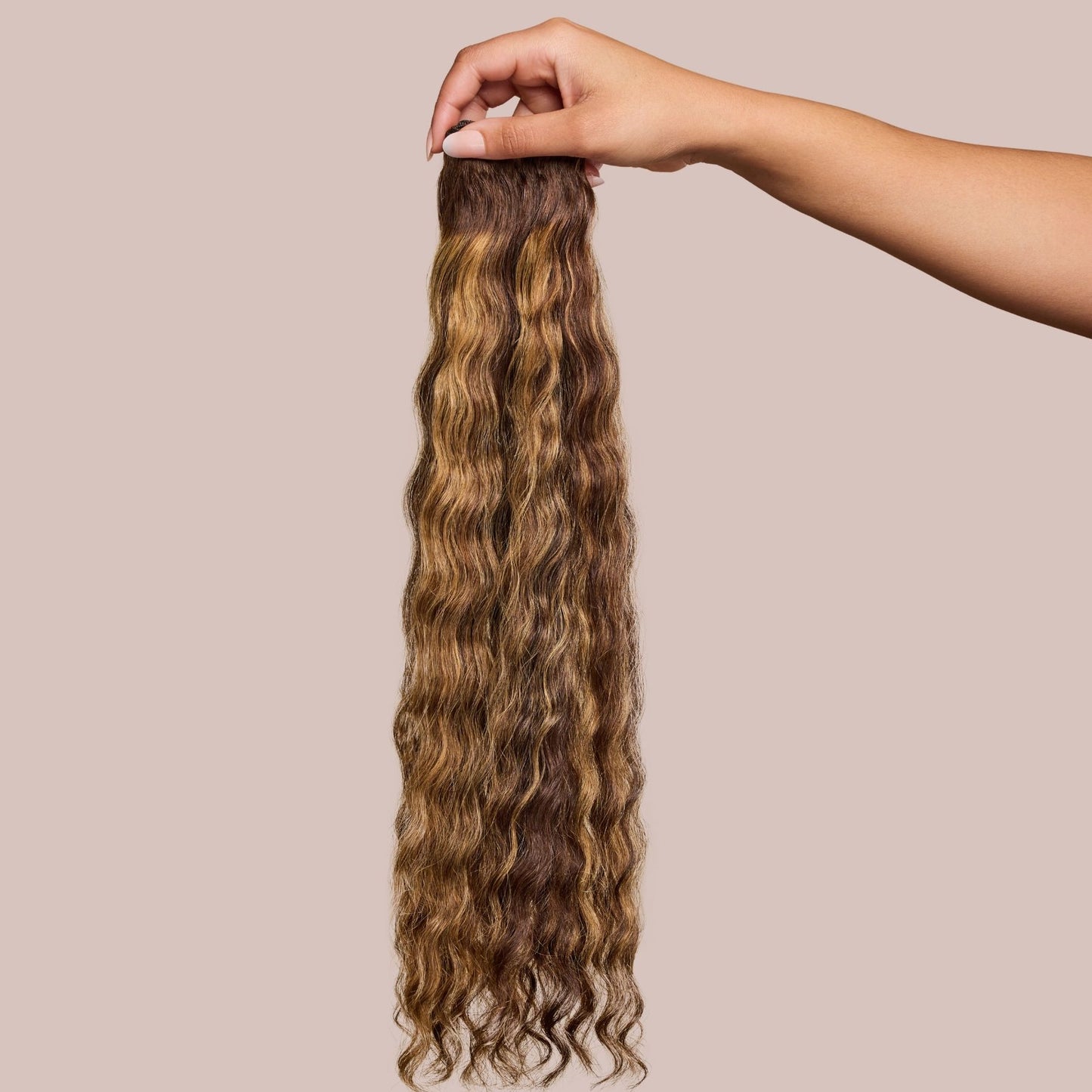 A hand delicately holding a bundle of luxury textured hair extensions in a rich, wavy pattern with a blend of warm brown and honey-blonde tones. The high-quality extensions have a natural shine and soft texture, perfect for adding volume and length to textured hairstyles.