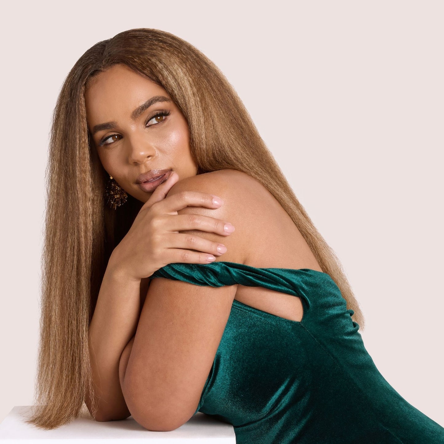 A glamorous woman with long, silky kinky straight hair extensions in a golden brown shade. Her voluminous, blown-out hair cascades naturally as she leans gracefully on a white pedestal, wearing an elegant emerald green velvet dress. These premier textured hair extensions add fullness, length, and a seamless blend, perfect for achieving a luxurious and sophisticated look.