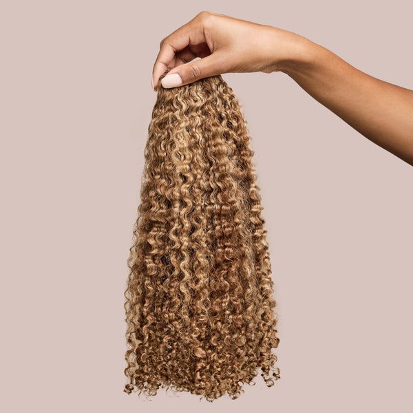 A hand holding a bundle of natural curly hair extensions in a stunning golden espresso shade. The well-defined curls are soft and bouncy, cascading naturally with deep brown tones infused with warm golden highlights, adding richness and dimension. These luxury textured hair extensions provide an effortless blend with natural curly hair, offering enhanced length, volume, and versatility for protective styling.