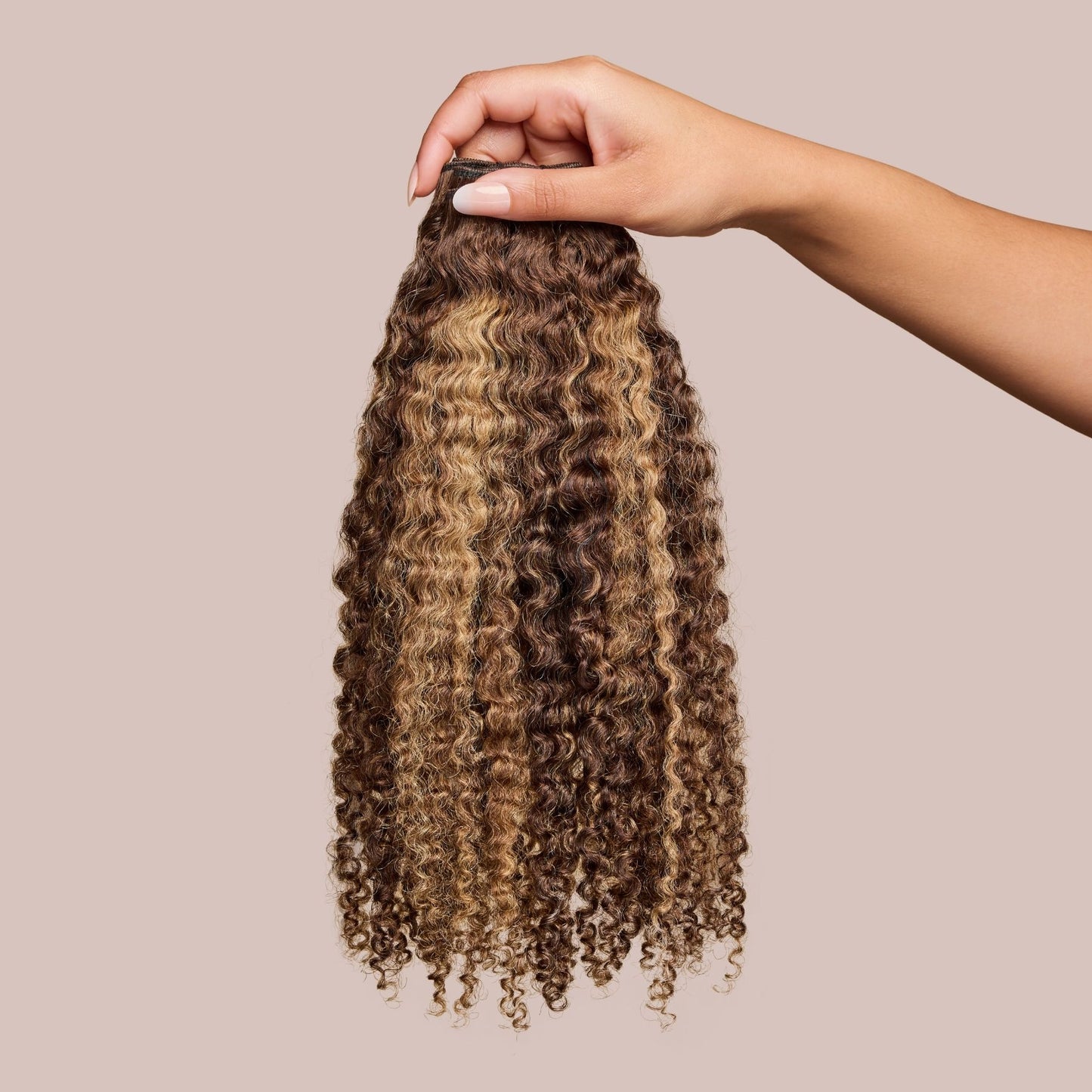 A hand elegantly holding a bundle of luxury textured hair extensions with tight, springy curls in a blend of deep brown and golden blonde highlights. The premium-quality extensions showcase a voluminous, natural texture, ideal for enhancing curly hairstyles with added length and dimension.