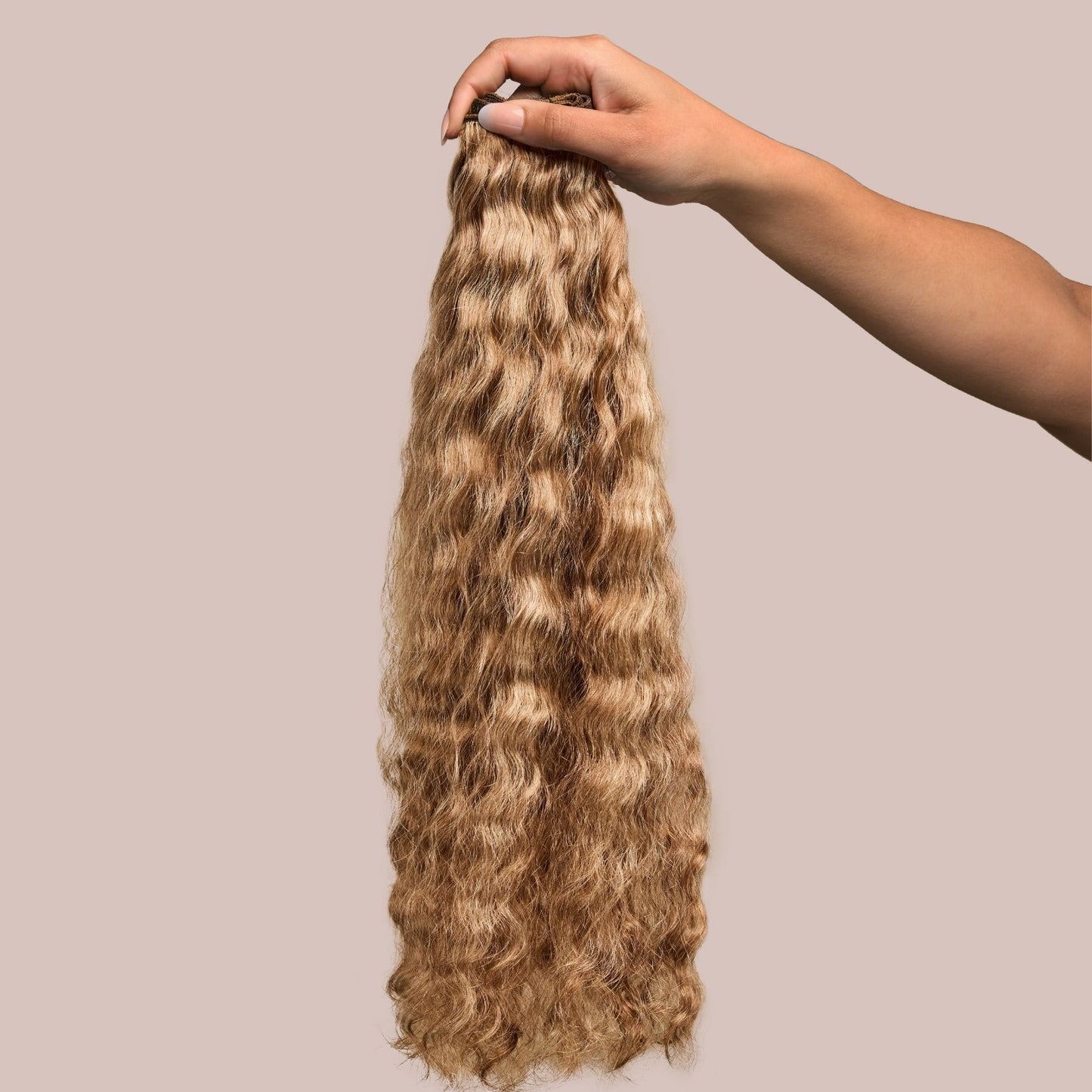 A hand holding a bundle of loose wavy textured hair extensions in a rich caramel shade. The soft, flowing waves have a silky texture with warm golden-brown tones, adding depth and dimension for a naturally radiant look. These premier textured hair extensions provide versatility for styling, making them an excellent choice for adding length, fullness, and a seamless blend with natural hair.