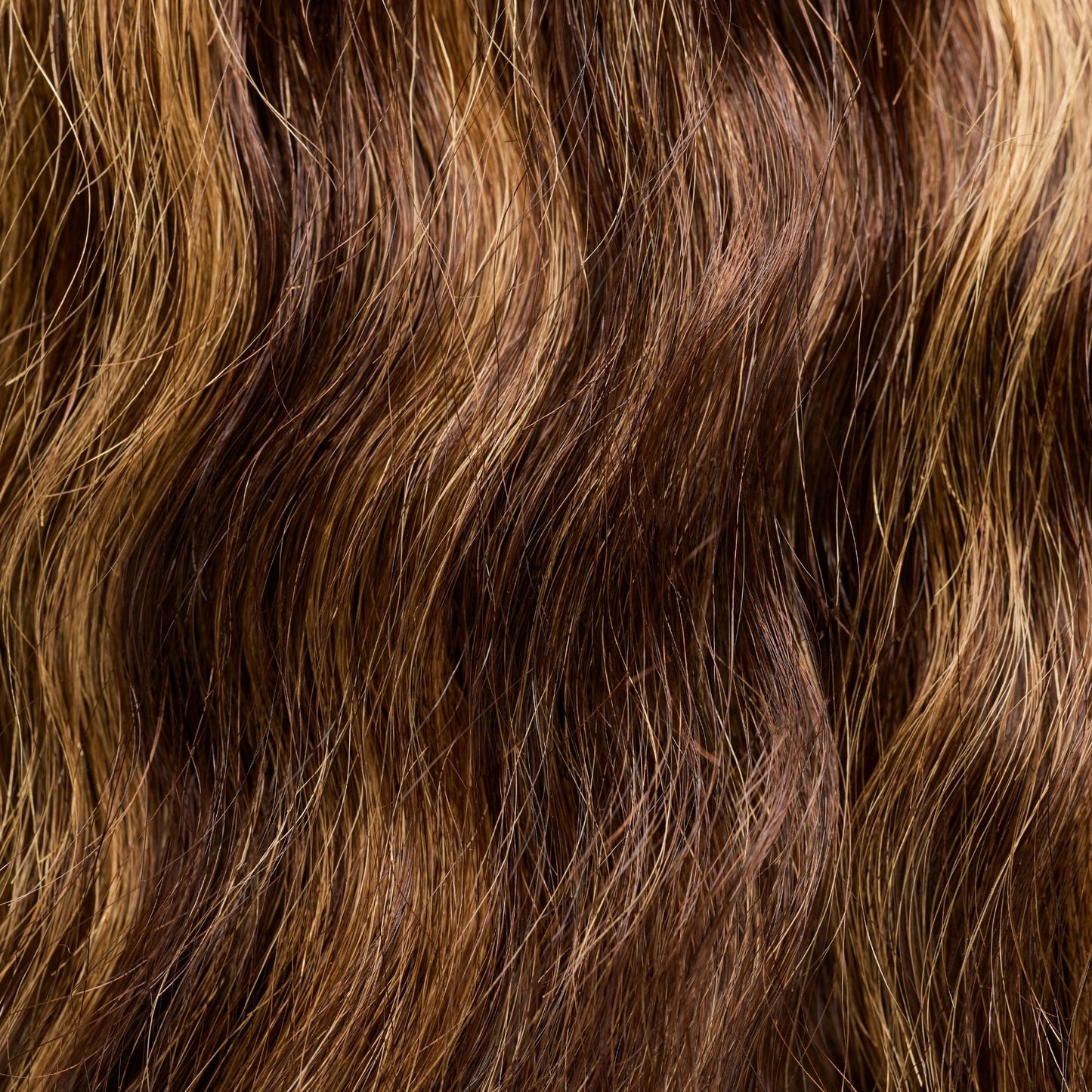 A close-up texture shot of loose wavy textured hair extensions in a rich brown shade with golden highlights. The soft, flowing waves add volume and dimension, creating a natural, effortless look. These premier textured hair extensions mimic the beauty of naturally wavy hair, providing a lightweight feel and a glossy, healthy sheen, making them ideal for versatile styling.