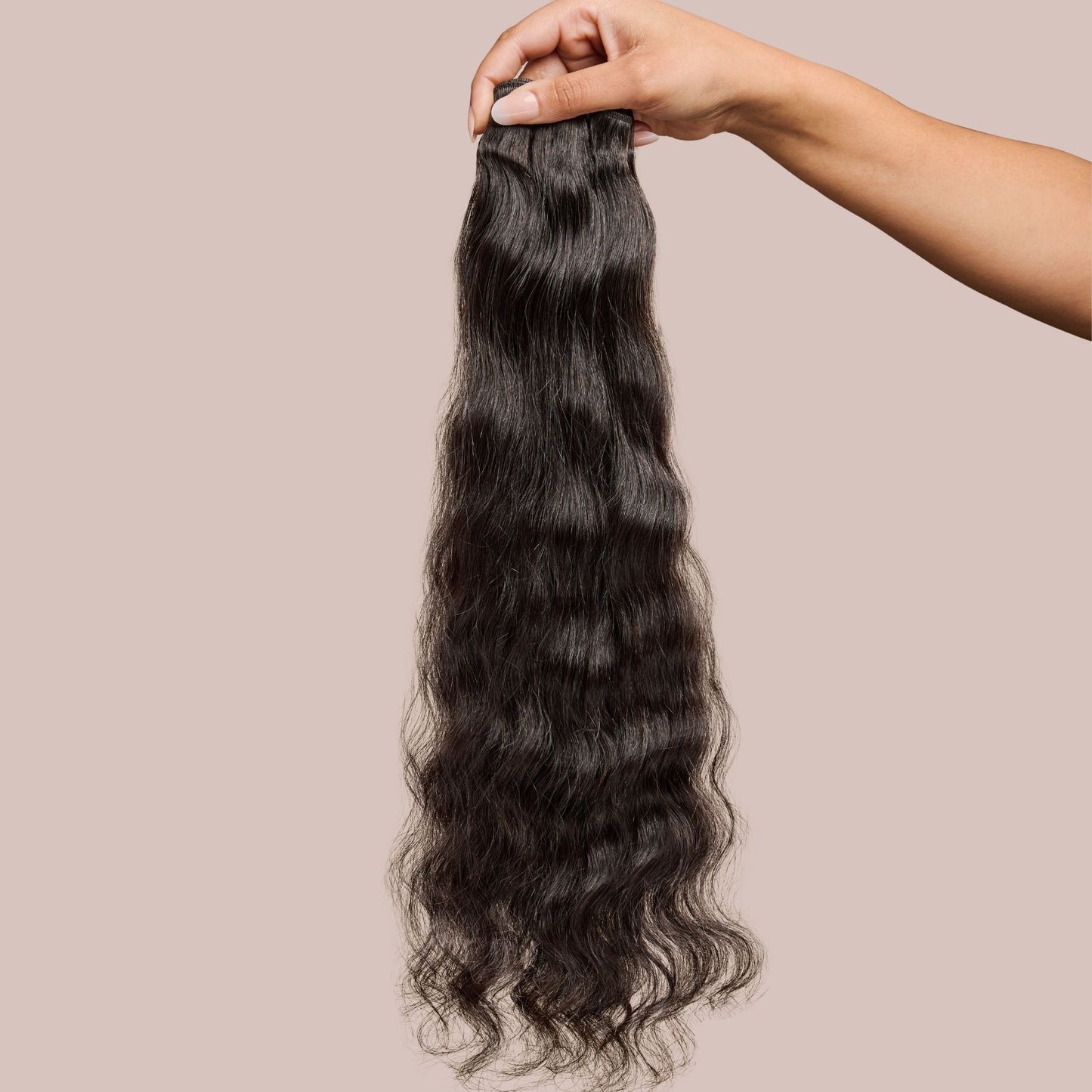 A hand holding a bundle of loose wavy textured hair extensions in a natural black shade. The soft, flowing waves have a silky texture with a subtle sheen, creating a voluminous and effortlessly glamorous look. These premier textured hair extensions provide versatility for styling, making them an excellent choice for adding length, fullness, and a seamless blend with natural hair.