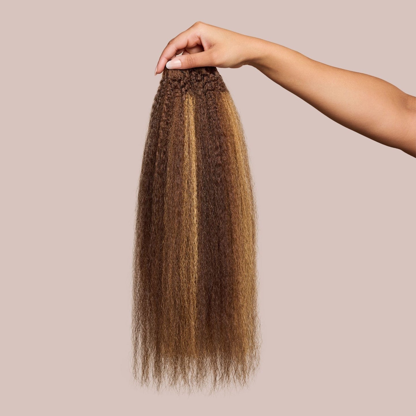 A hand holds a bundle of kinky straight hair extensions in a rich brown shade with golden blonde highlights, showcasing the soft yet voluminous texture. These luxury textured hair extensions mimic natural blow-dried hair, providing fullness and versatility for seamless blending with natural hair. The high-quality strands offer a realistic, lightweight feel, making them ideal for protective styling and enhancing volume effortlessly.